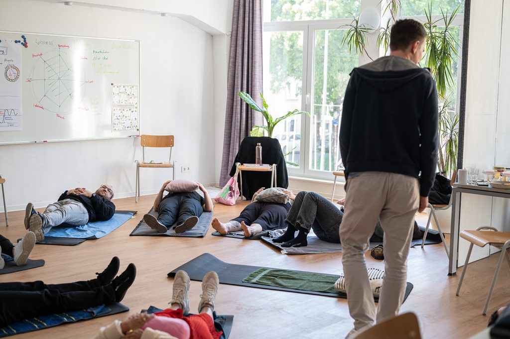Mehrere Personen liegen auf Yogamatten mit Kissen auf ihren Bäuchen. Ein Trainer beobachtet die Gruppe.